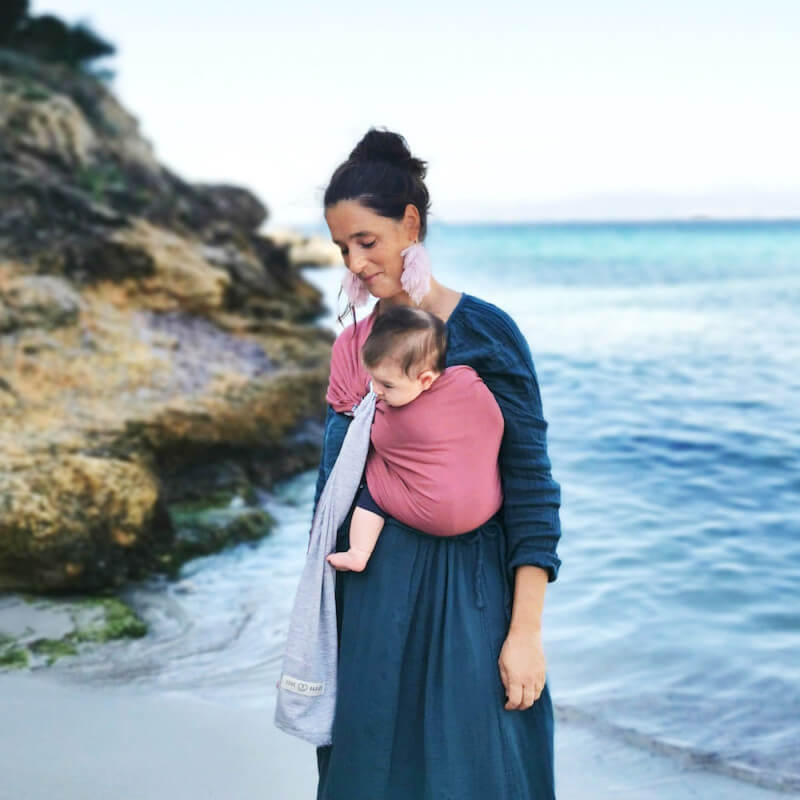 Mutter trägt Baby in Love Radius Sling in Rosa auf felsigem Strand