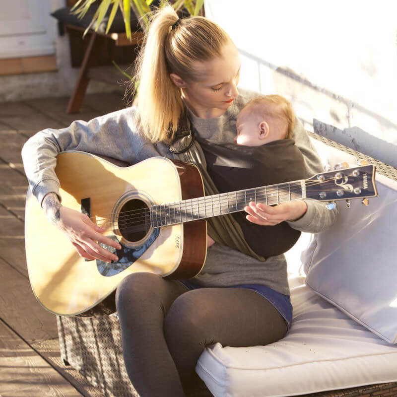 Kvinna spelar akustisk gitarr med baby i sling Love Radius antracit-oliv reversibelt