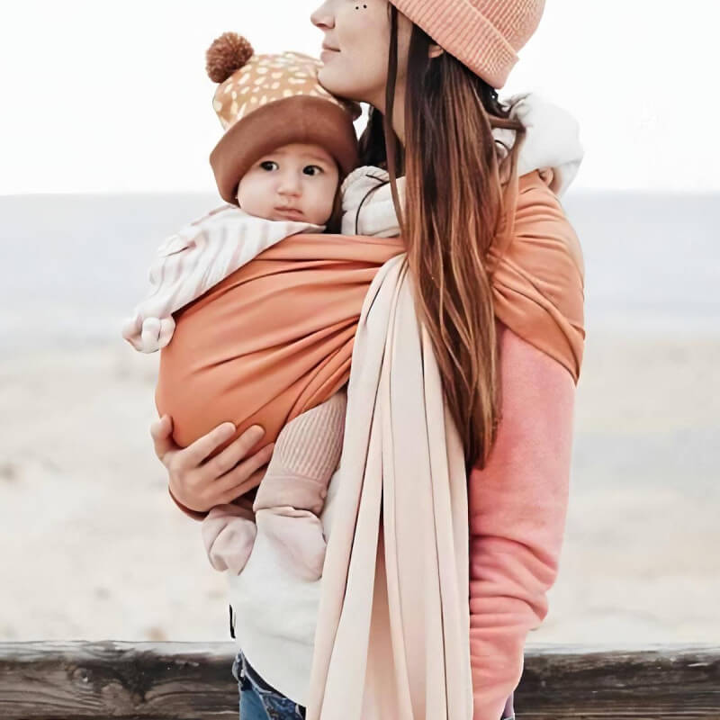 Mãe carregando bebê em Love Radius Sling reversível nude caramelo na praia