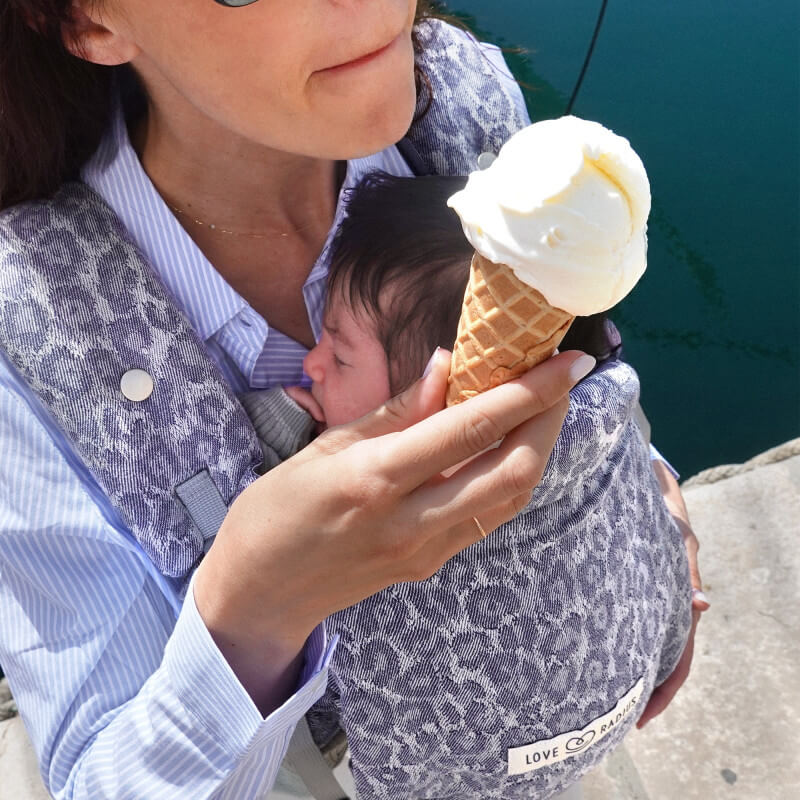 Frau trägt Baby in Love Radius PhysioCarrier 2 Blue Leopard Babytrage am Meer und genießt Eis