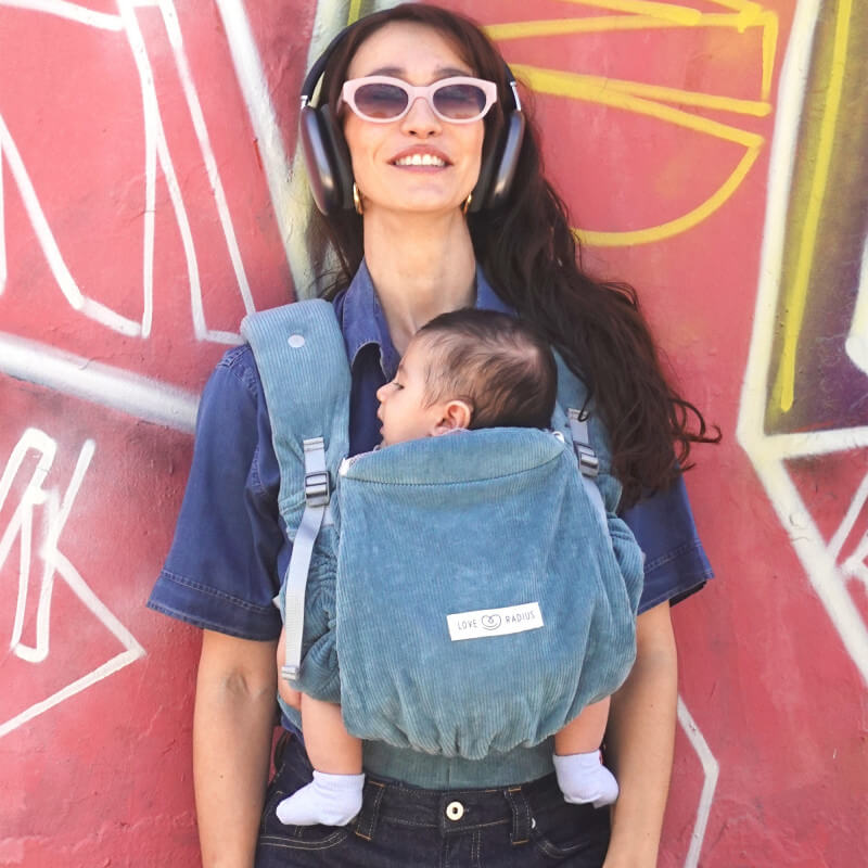 Femme portant le porte-bébé Love Radius PhysioCarrier 2 Velvet Blue avec bébé, fond mur coloré