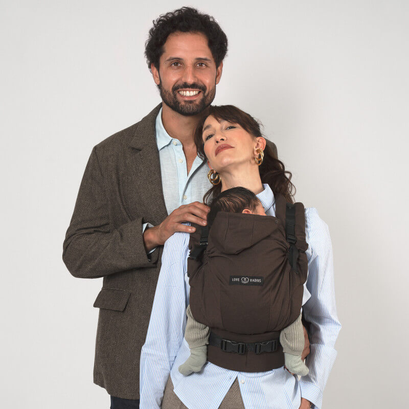 Mujer llevando Love Radius HoodieCarrier 2 Wood, bebé visible en posición frontal, hombre sonriendo detrás